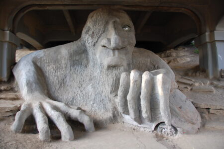 A large sculpture of a troll with a giant hand is partially buried under a bridge. The troll has detailed facial features and is grasping a real car with one hand. The setting includes rocky terrain and the underside of the bridge.