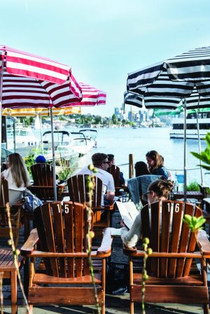 People sitting on wooden chairs with striped umbrellas overhead, overlooking a waterfront with boats and a city skyline in the background. The setting appears relaxed and sunny.