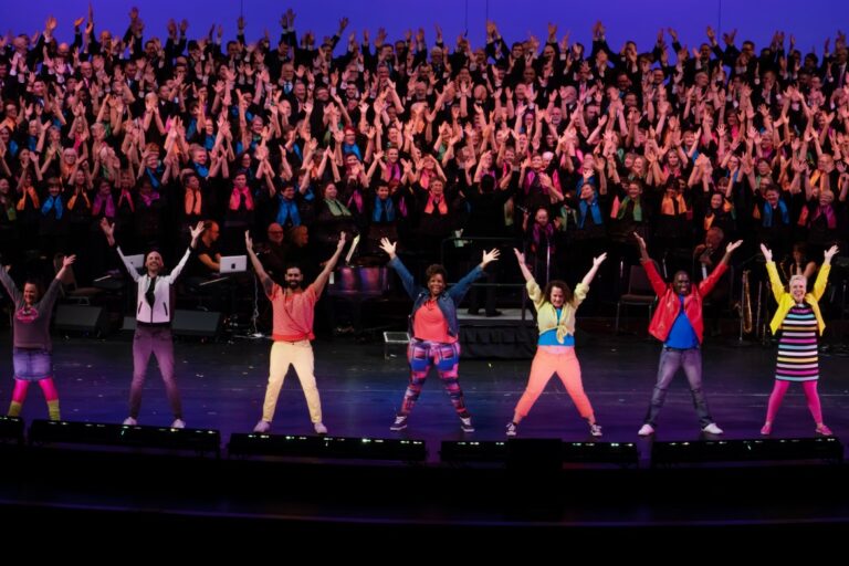 A group of diverse performers in colorful outfits stands on stage with arms raised. A large choir in coordinated attire sings in the background under blue and purple lighting.
