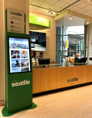 Seattle visitor center desk with green signage and digital screen displaying attractions. An information board is visible in the background. A staff member stands behind the counter ready to assist visitors.