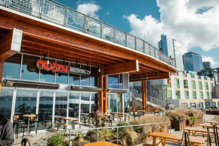 Outdoor brewery with wooden accents on a sunny day, featuring the sign "Old Stove Brewing Co." The patio has tables and chairs, with city buildings and partly cloudy skies in the background.