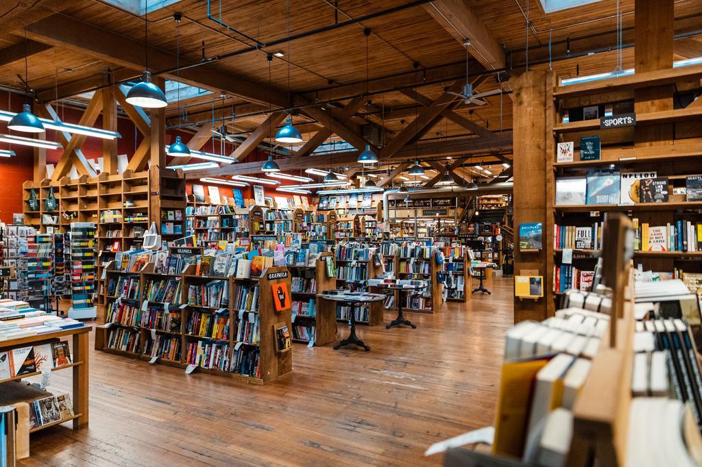 A spacious, well-lit bookstore with wooden floors and beams, filled with shelves and tables displaying a variety of books. Blue pendant lights hang from the ceiling, creating a warm and inviting atmosphere.