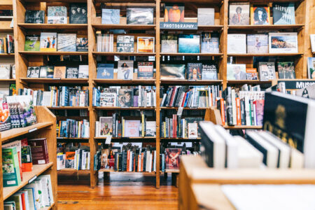 A cozy bookstore scene features tall wooden shelves filled with various books. The foreground has books displayed on a table, while the background shelves include books on photography and other genres, creating a warm and inviting atmosphere.