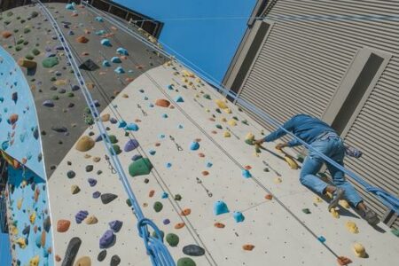A person climbs an outdoor rock wall with multicolored holds, wearing jeans and a blue long-sleeve shirt. Blue ropes hang down the wall. The building and clear sky are visible in the background.