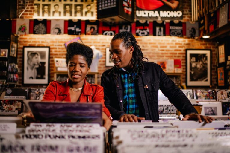Two people browse records in a cozy vinyl store with a brick wall and music-themed decor. The woman wears a red jacket, and the man wears a black jacket over a plaid shirt. Shelves are filled with records and posters on the walls.