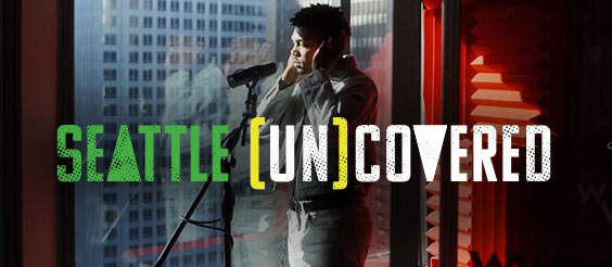 A person sings into a microphone in a studio with city skyscraper views. The text "Seattle [Un]covered" is prominently displayed over the image.