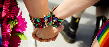 Two people holding hands. One person wears a colorful jeweled bracelet. Nearby is a bouquet of pink flowers and a rainbow-striped item, possibly a flag. The background shows a blurred sidewalk and a person's leg in black clothing.