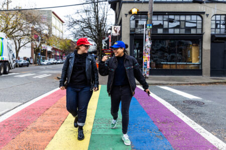 DJ Chong the Nomad, wearing all black with a blue ball cap, walks her friend, Jax, wearing all black with a red ball cap, across a rainbow crosswalk in Capitol Hill.