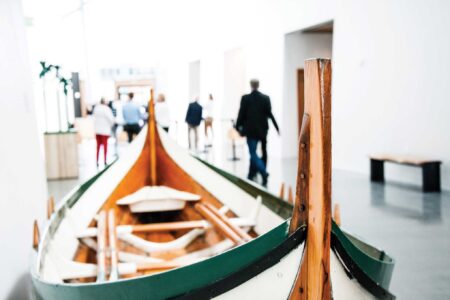 A wooden boat with green accents is displayed indoors in a bright gallery space, reminiscent of the modern, minimalistic vibe you’d find in Seattle's Ballard neighborhood. In the distance, a group of people walks by, slightly out of focus.