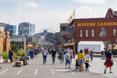 People walk along a busy waterfront boardwalk lined with shops, restaurants, and colorful signs. The Miners Landing building is visible, with city buildings and a stadium arch in the background on a sunny day.