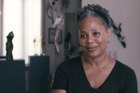 A person with short curly hair and a black shirt smiles softly while sitting indoors. The background is softly lit with sculptures and artworks visible, creating an artistic and relaxed atmosphere.
