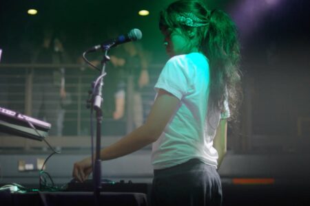 A person with long dark hair and a bandana is performing at a dimly lit venue. They are wearing a white t-shirt, standing in front of a microphone and DJ equipment, with an elevated audience area in the background.