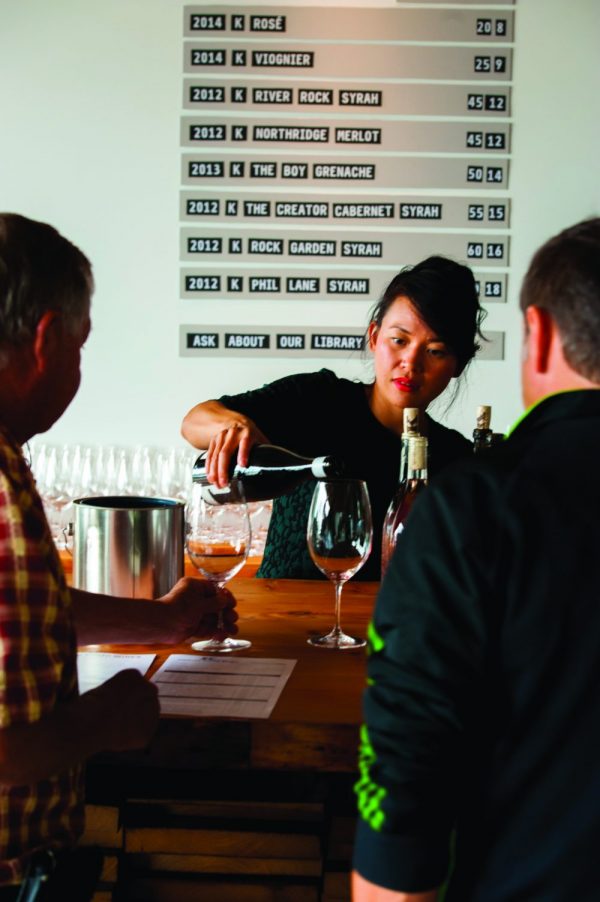 A woman pours wine into a glass for a customer at a tasting event. Behind her, a chart on the wall lists various wines with their details. Two men are at the counter observing the pouring.