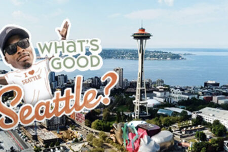 Aerial view of Seattle with a large text overlay reading "What's Good Seattle?" A person wearing a cap and sunglasses is also part of the overlay, appearing excited and welcoming. The Space Needle is prominently visible in the background.
