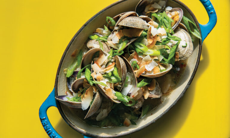 A blue-handled ceramic dish filled with clams, garnished with sliced green onions and toasted coconut flakes, on a bright yellow background.