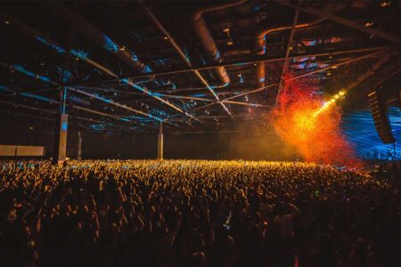 A large, energetic crowd at an indoor concert venue, with colorful lights and confetti falling from above. A stage is visible in the distance, illuminated by bright spotlights. The audience has raised hands, enjoying the lively atmosphere.