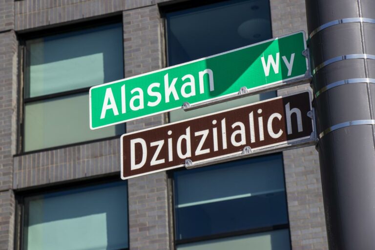 Street signs on a pole with "Alaskan Way" in green and "Dzidzilalich" in brown, set against a background of a brick building.