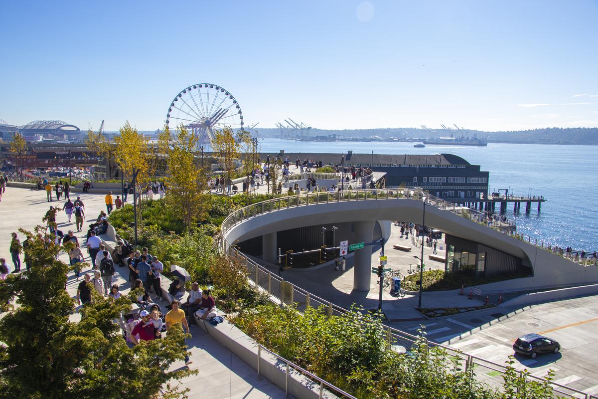 People stroll along a sunny waterfront park with a large Ferris wheel, green spaces, and views of the water and industrial cranes in the background. The sky is clear and blue.