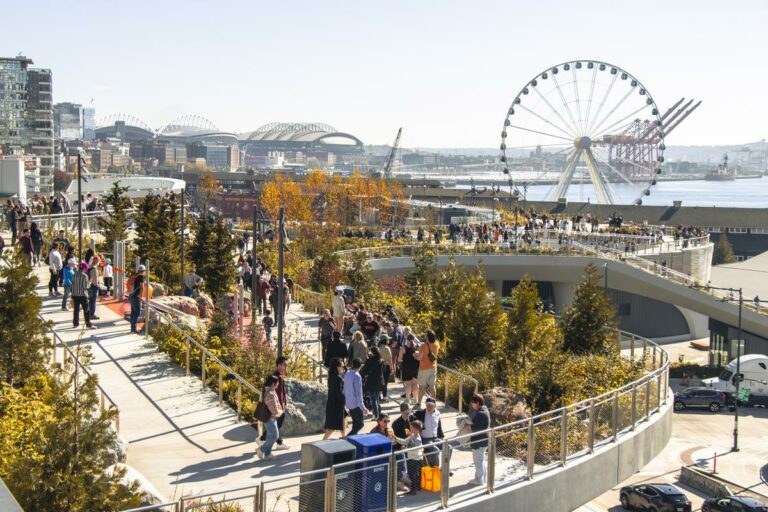 A bustling urban park features people walking along paths surrounded by greenery. In the background, a large Ferris wheel overlooks a waterfront, with bridges and city skyline visible on a clear, sunny day.