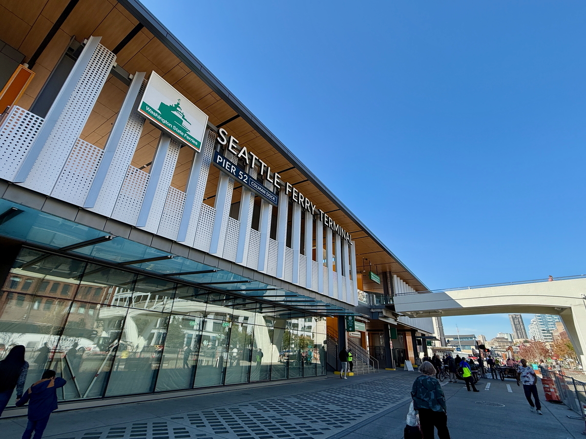 People walk outside the modern Seattle Ferry Terminal at Pier 52 on a clear, sunny day. The building’s sign and glass entrance are visible, with city buildings and a blue sky in the background.