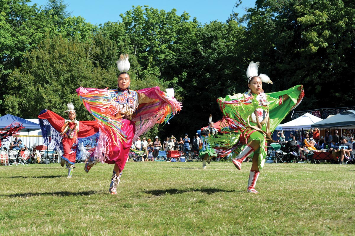 Dancers in vibrant traditional attire perform at an outdoor event. The colorful outfits feature fringe and beadwork, and the dancers are mid-motion on a grassy field. A crowd watches in the background under trees and tents.