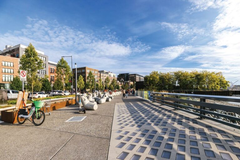A scenic urban pathway on a sunny day, featuring modern buildings, trees, and a row of abstract sculptures. People walk and bike along the path, bordered by a railing overlooking the water. A green rental bike is parked on the side.