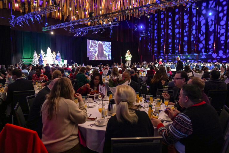 A large indoor holiday event with numerous people seated at round tables, dining and watching a stage presentation. The venue is decorated with illuminated trees and colorful hanging lights, creating a festive atmosphere.