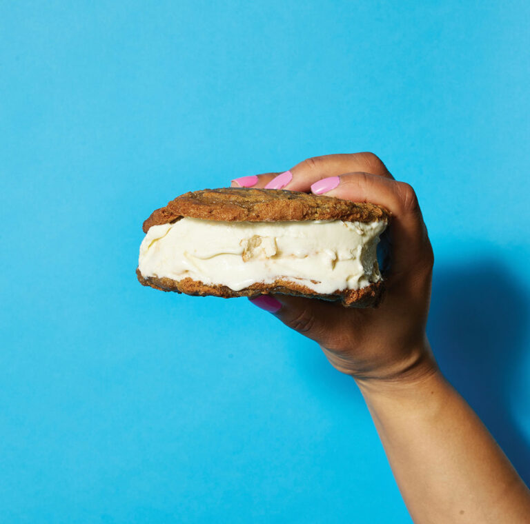 A hand with pink-painted nails holds a chocolate chip cookie ice cream sandwich against a bright blue background. The sandwich features vanilla ice cream between two large cookies.