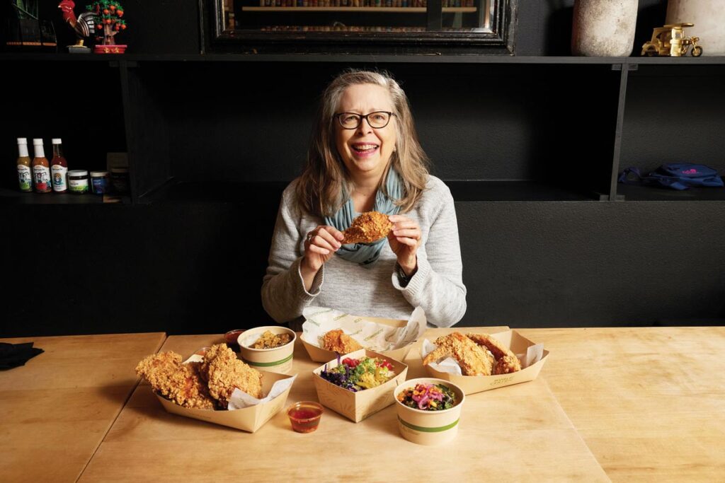 A woman with long hair and glasses smiles while holding a piece of fried chicken. She sits at a table with various dishes, including more fried chicken and colorful side dishes, on a wooden tabletop. Shelving with items is visible in the background.
