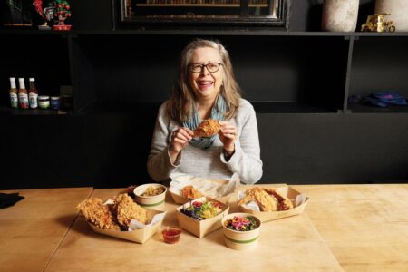 A woman with long hair and glasses smiles while holding a piece of fried chicken. She sits at a table with various dishes, including more fried chicken and colorful side dishes, on a wooden tabletop. Shelving with items is visible in the background.