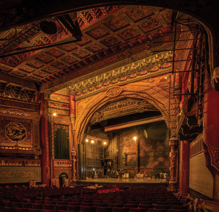 Experience the opulence of a Seattle theater, where an ornate interior boasts a richly decorated ceiling, intricate carvings, and a grand proscenium arch framing the empty stage. Rows of plush red seats fill the foreground, while warm lighting casts a golden glow—one of many captivating things to do in Seattle.