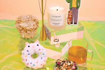 A colorful still life featuring a candle, a decorative figurine, a cup of tea, a beaded bracelet, a donut-shaped object with sprinkles, a cluster of translucent crystals, and a brass necklace stand on a vibrant green and peach background.
