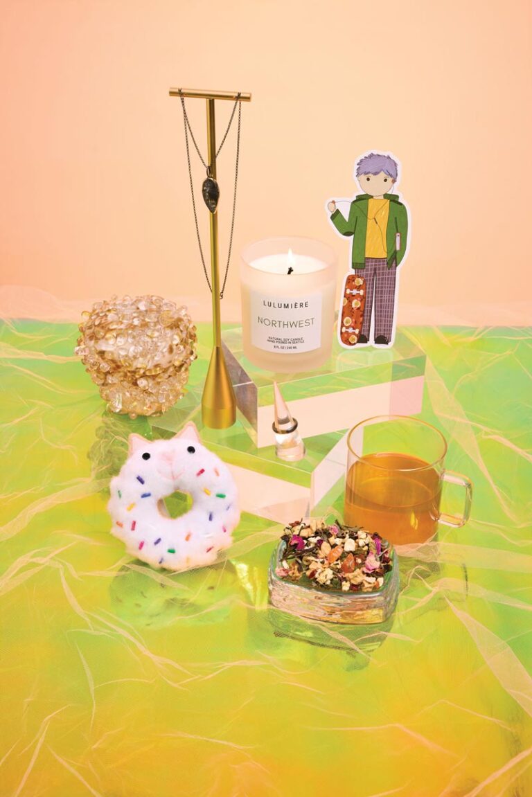 A colorful still life featuring a candle, a decorative figurine, a cup of tea, a beaded bracelet, a donut-shaped object with sprinkles, a cluster of translucent crystals, and a brass necklace stand on a vibrant green and peach background.