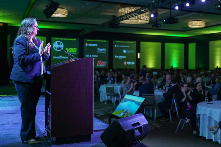 A woman in a suit stands at a podium, clapping, in a large green-lit conference room filled with seated attendees. Logos are projected on a screen in the background.