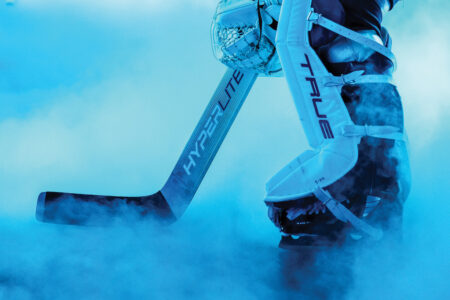 Close-up of a hockey player's legs in goalie gear, with a thick cloud of mist or fog surrounding them. The player holds a hockey stick labeled "HYPERLITE" and wears protective pads labeled "TRUE." The scene is illuminated with a blue tint.