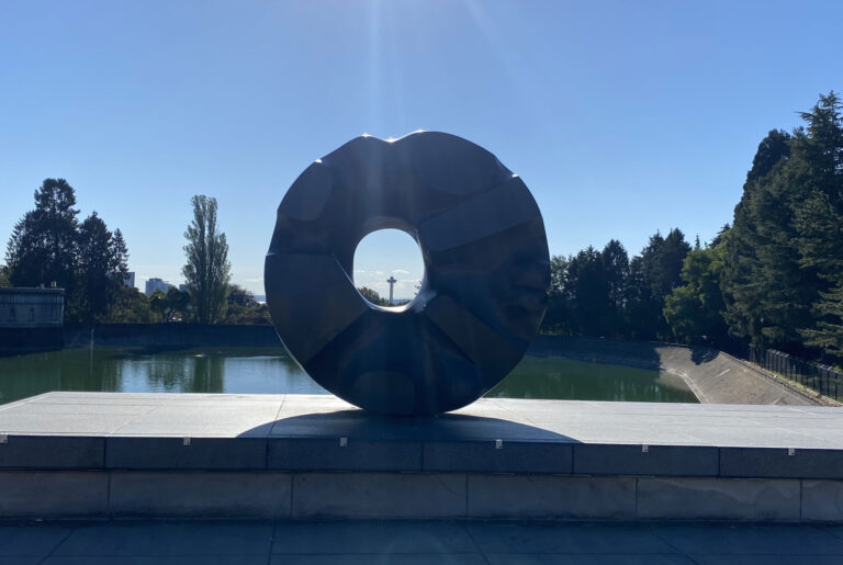 A large, dark circular sculpture with a hollow center stands on a platform—a serene spot among the top things to do in Seattle. Behind it, a calm body of water is surrounded by trees under a clear blue sky, where sunlight creates a halo effect around the sculpture.