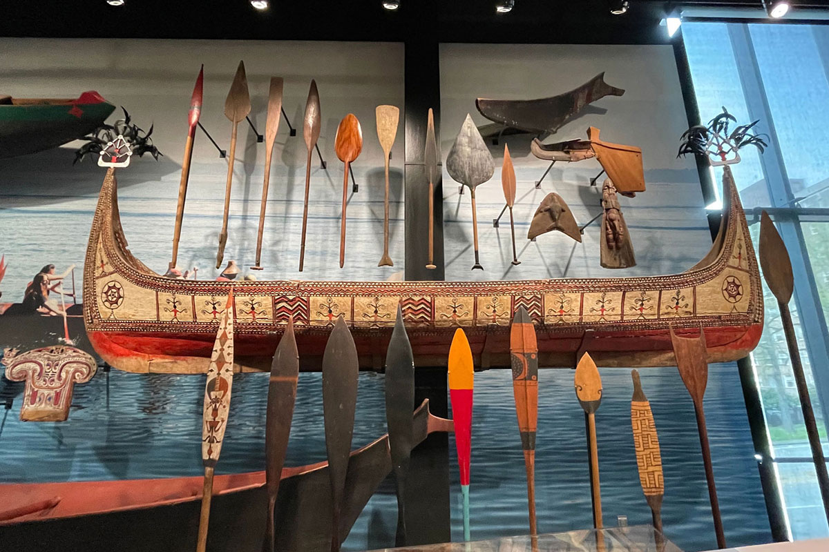 A traditional outrigger canoe with intricate designs is displayed on a wall, surrounded by an array of paddles and canoe decorations. The background depicts a serene water scene, enhancing the cultural and historical ambiance.
