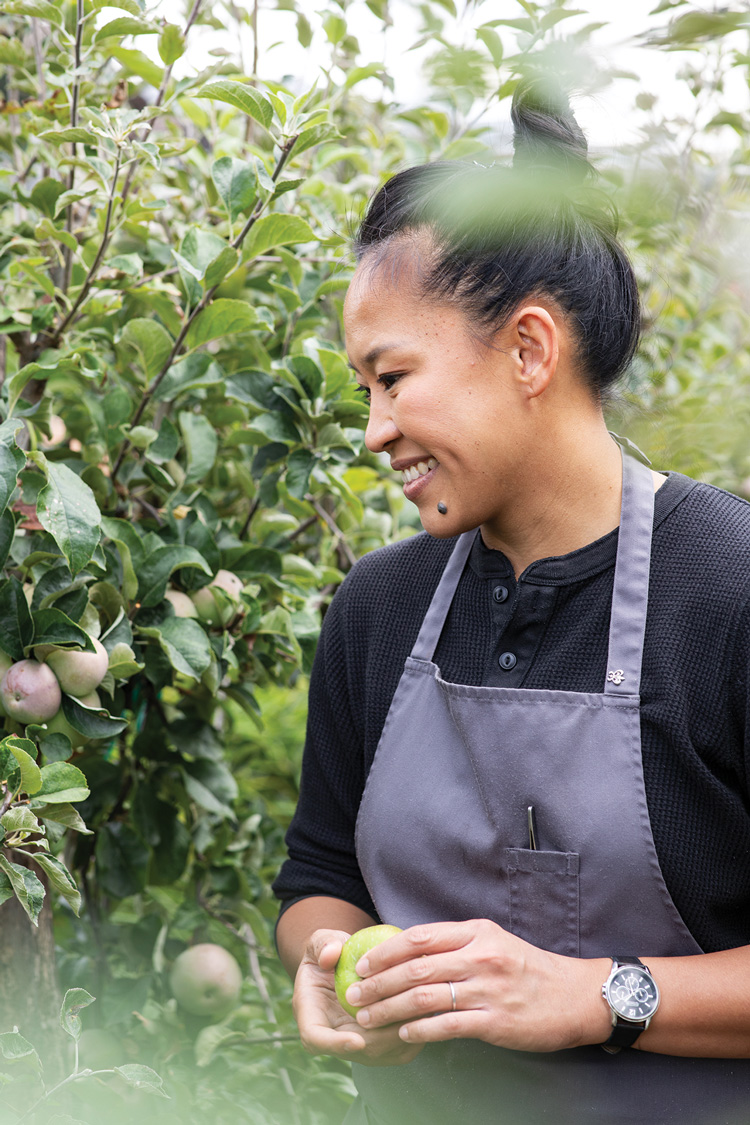 A person is smiling and holding a green apple in an orchard. They are wearing a dark shirt and a gray apron, with leafy apple trees in the background. The atmosphere is peaceful and natural.