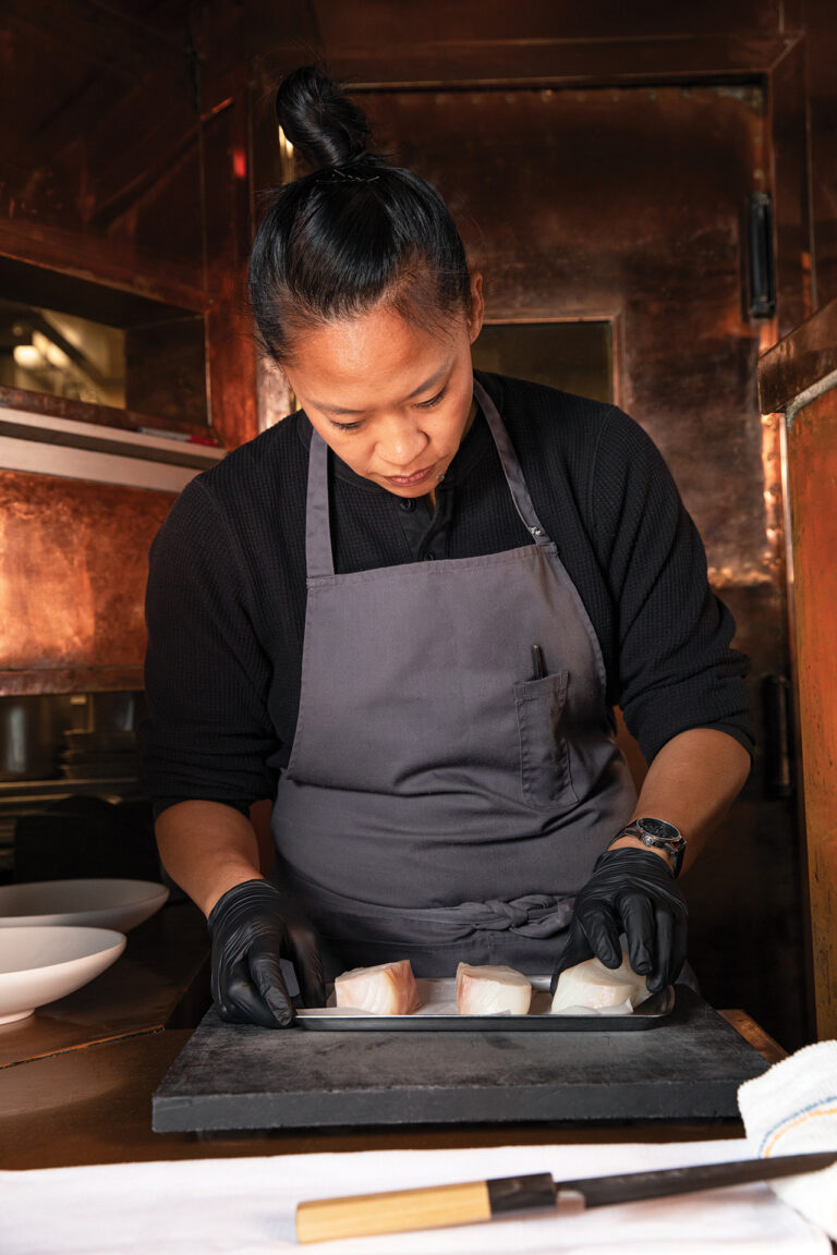 A person wearing a gray apron and black gloves is carefully preparing or arranging fish fillets on a black tray in a kitchen. The background features copper-colored surfaces and kitchen equipment.