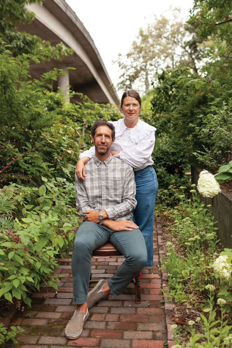 A man sits on a chair while a woman stands beside him, resting her hand on his shoulder. They are surrounded by lush greenery and a brick pathway, with an overpass visible in the background. Both are casually dressed and smiling.