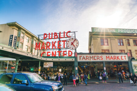 People walking on a cobblestone street in front of a building with a large neon red sign that reads Public Market Center.