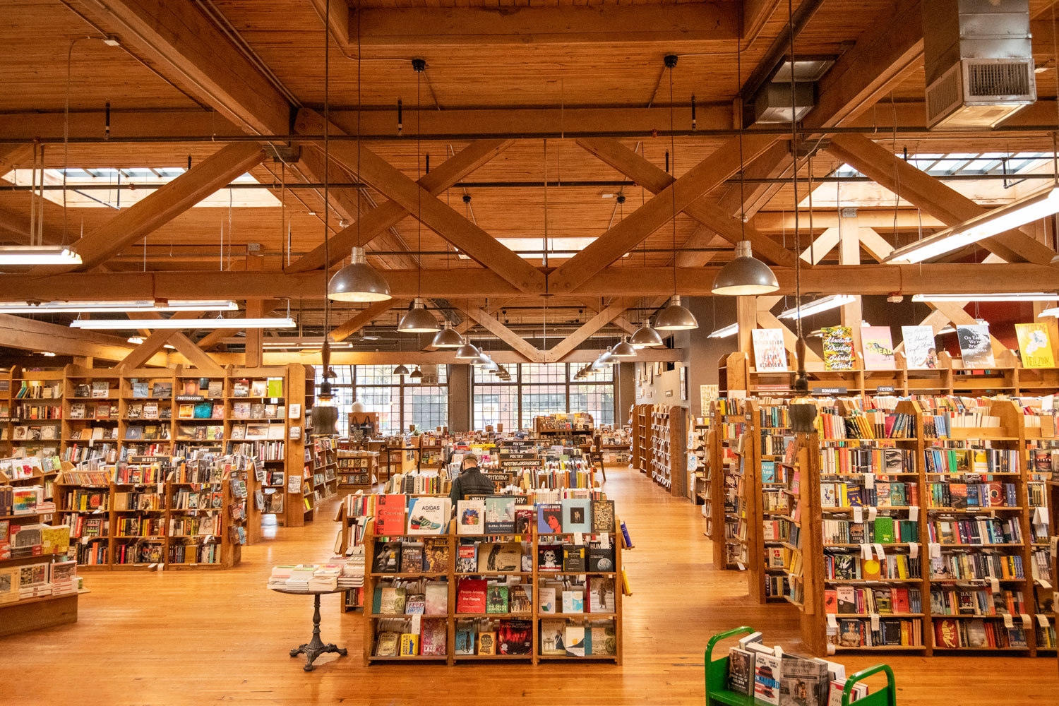 A spacious bookstore with rows of wooden bookshelves filled with various books. The ceiling features exposed wooden beams and modern lighting. Large windows allow natural light to illuminate the space.