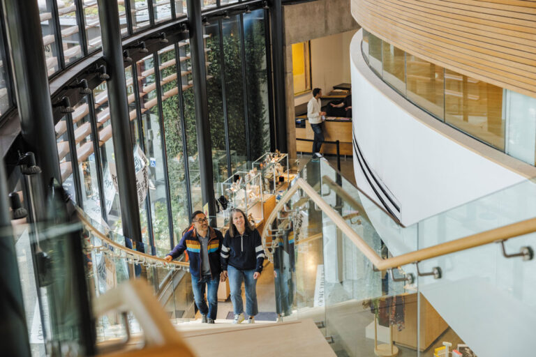 Two people walk up a modern, curved staircase in a bright building with large glass windows—evoking the airy style seen in Bainbridge Island and Bremerton—while another person stands near a counter. The space features wood accents and natural light.