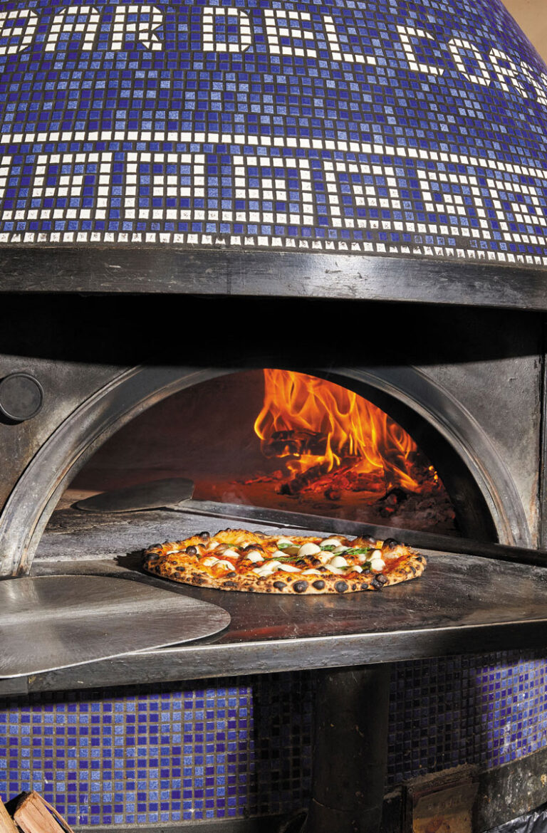 A wood-fired pizza oven with blue mosaic tiles and visible flames baking a freshly prepared pizza on a metal peel inside.