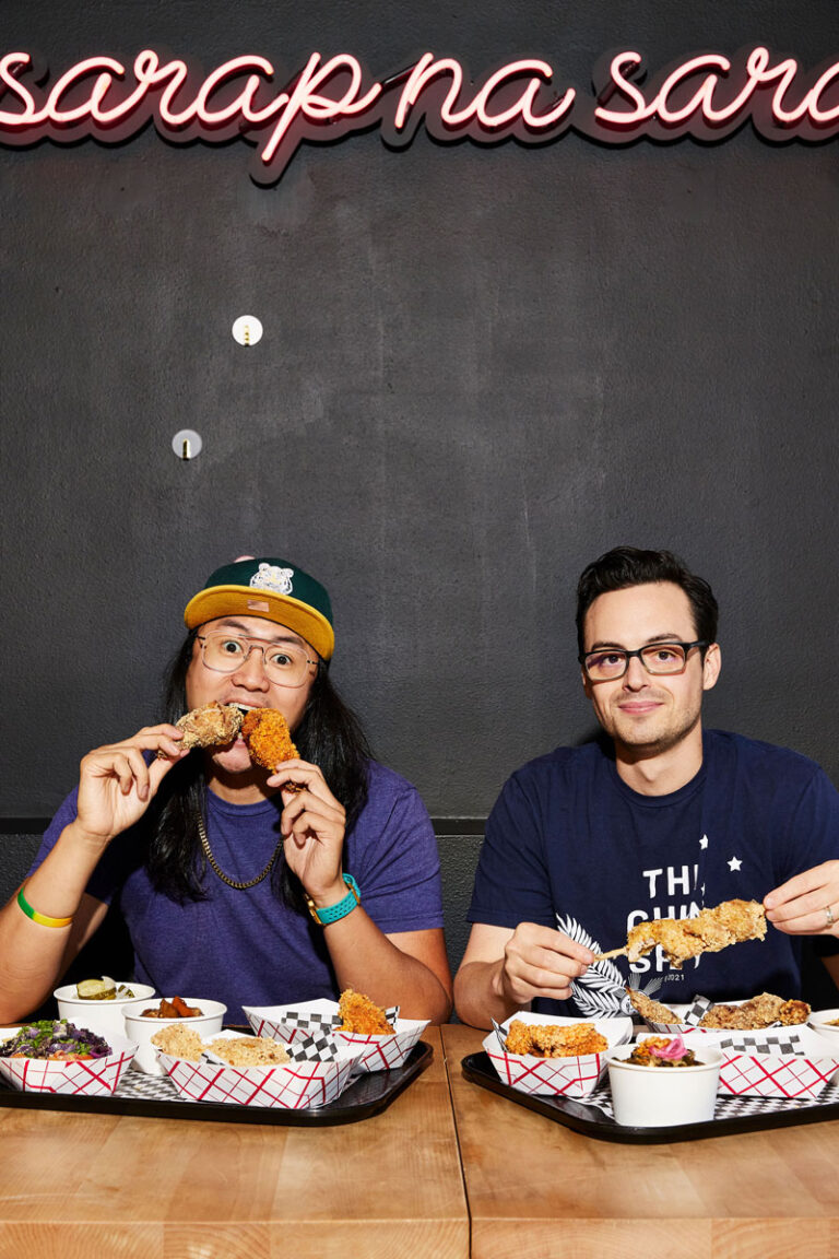 Two men sit at a table in one of the North Seattle Neighborhoods, enjoying trays of fried chicken and sides. One man, wearing glasses and a cap, bites into a drumstick as the other smiles and holds up his chicken under a neon sign.