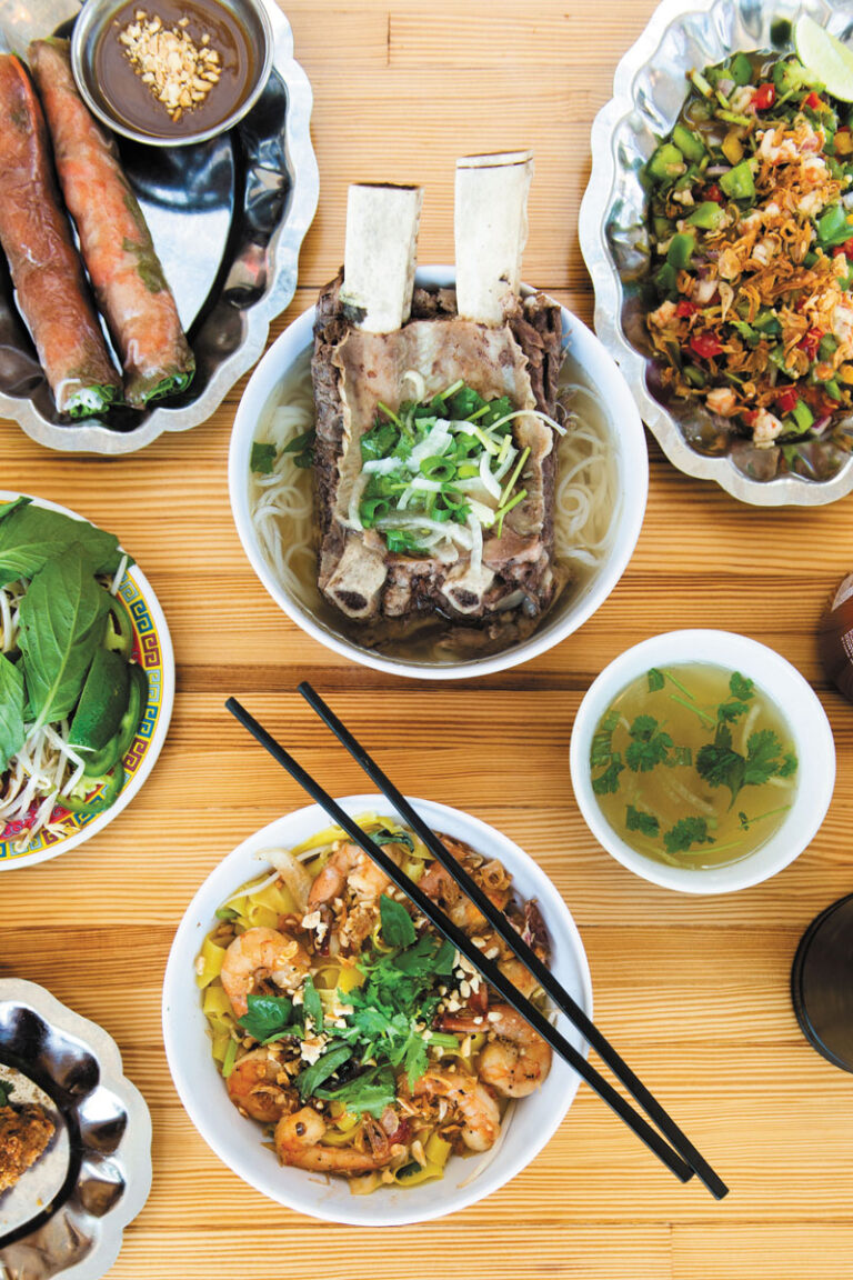 A table set in Chinatown-International District with Vietnamese dishes: a bowl of noodle soup with beef ribs, spring rolls, noodles topped with herbs and peanuts, a side of broth, and a plate of stir-fried vegetables.