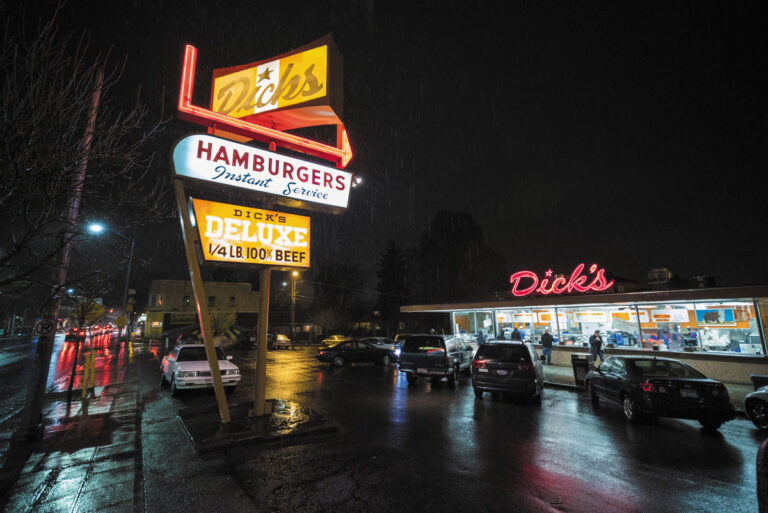 A retro-style Dick's Hamburgers diner at night sits at Fremont & Wallingford, neon signs glowing and cars parked outside. People are visible inside the bright diner, with wet streets and pavement shining from recent rain.