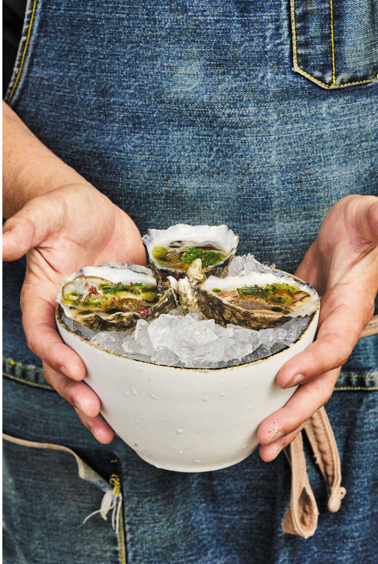 In the West Seattle neighborhood, a person wearing a denim apron holds a white bowl filled with ice and three opened oysters, garnished with herbs.