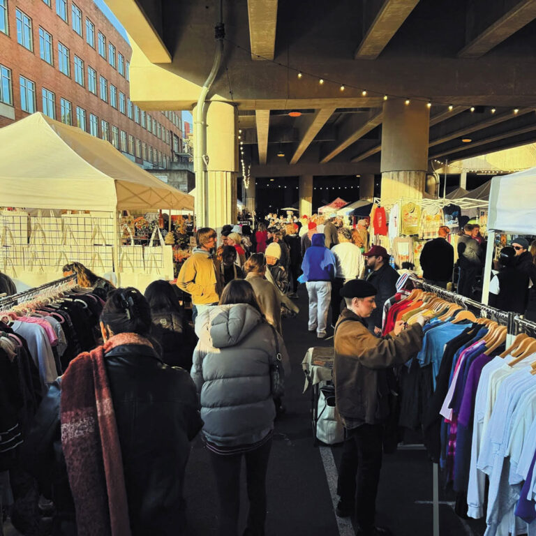 A busy outdoor market under a highway overpass in Fremont & Wallingford, with people browsing racks of clothing and various vendor tents set up. String lights hang overhead, and a brick building is visible in the background.