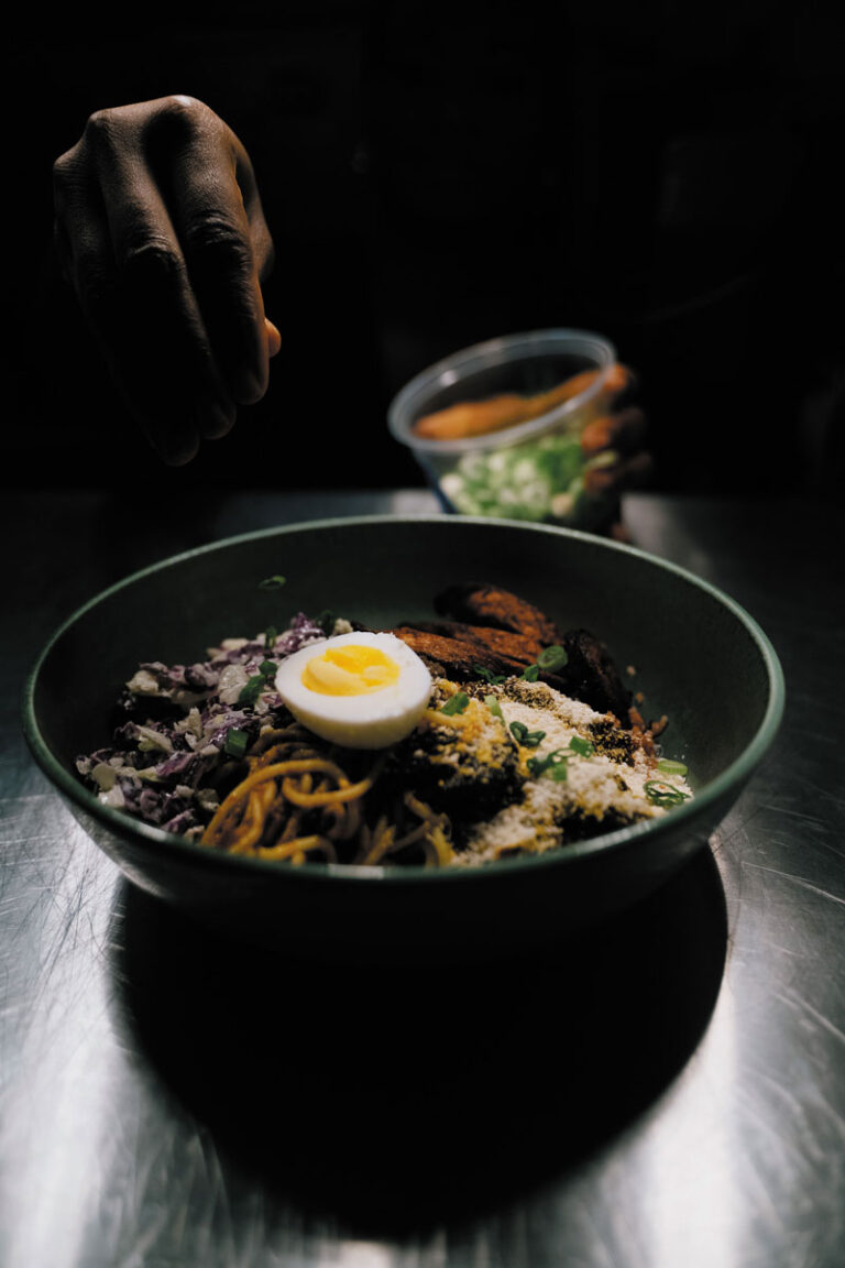 A hand sprinkles chopped green onions onto a bowl of noodles, vegetables, shredded purple cabbage, and a halved boiled egg—capturing the vibrant dining scene found in Seattle’s Capitol Hill neighborhood.